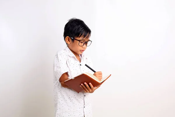 本に書いている間に立っているスマートなアジア系の少年 コピースペースと白い背景に隔離されています — ストック写真