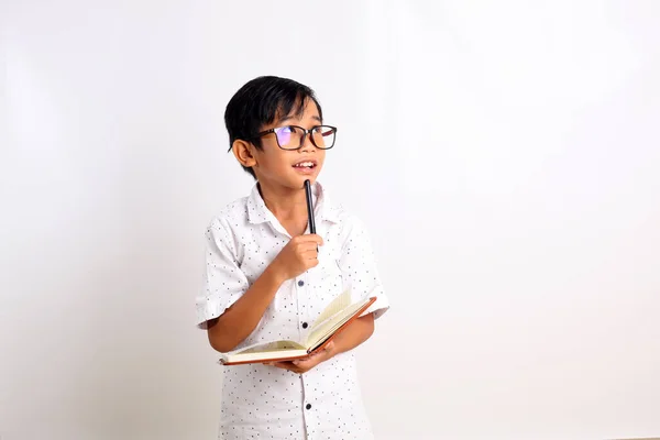 Happy asian schoolboy thinking while holding a book and looking sideways. Isolated on white background