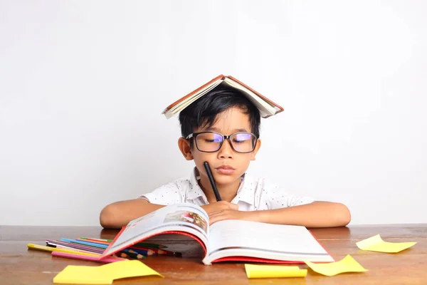 頭の上に本を置いて教室で勉強しているアジア系の少年のことを考えた 白地に隔離された — ストック写真