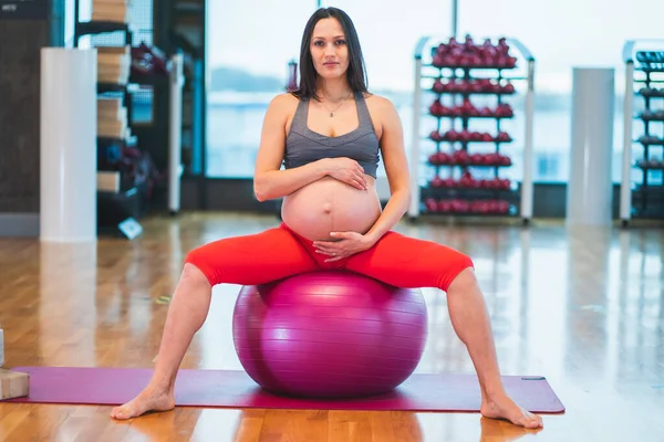 Full length portrait of pregnant woman sitting on fit-ball in gym