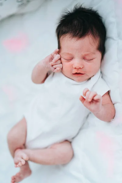 Amazing newborn baby with short dark hair in white bodysuit sleeping on back stirring in dream on white blanket.Vertical