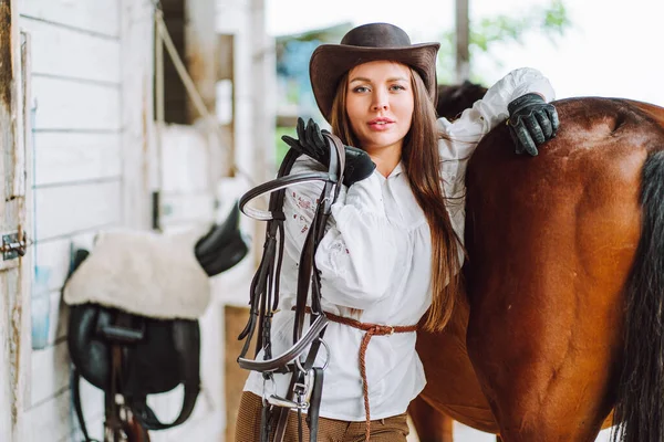 Confident horse woman cowgirl equestrienne hat, shirt, belt. Caring animal. Holding harness. Horseback riding