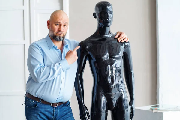 Bald serious adult man with beard, holding shoulders naked plastic black manikin