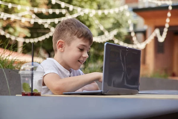 カフェのベランダにあるテーブルに座ってノートパソコンで勉強している白人の少年を笑顔にして — ストック写真