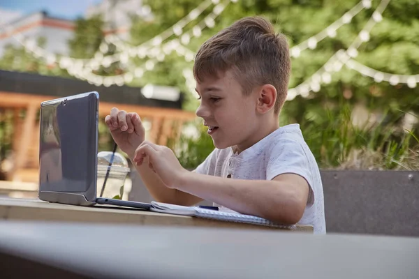 カフェのベランダにあるテーブルに座ってノートパソコンで勉強している白人の少年を笑顔にして — ストック写真