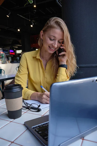 Νεαρή καυασιάτισσα όμορφη γυναίκα κάθεται με φορητό υπολογιστή στο καφέ, καλώντας από το smartphone — Φωτογραφία Αρχείου