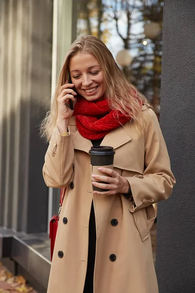 Kaukasisk kvinna i beige trenchcoat ringer via smartphone med kopp kaffe — Stockfoto