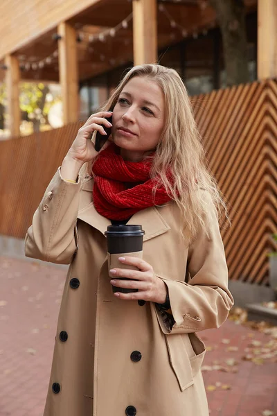 ベージュトレンチコートを着た白人女性がコーヒーを片手にスマートフォンを呼び — ストック写真
