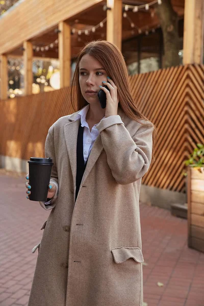 ベージュトレンチコートを着た白人女性がコーヒーを片手にスマートフォンを呼び — ストック写真