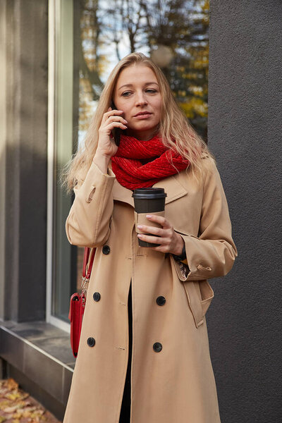 caucasian woman in beige trench coat calling by smartphone with cup of coffee