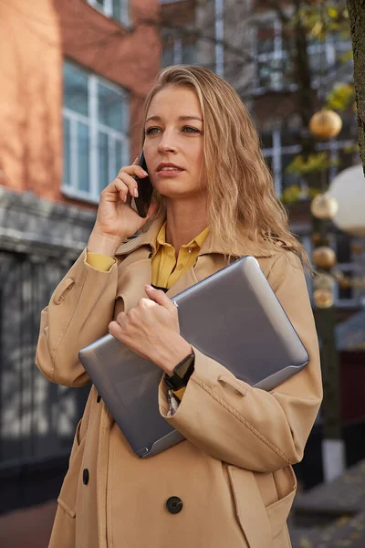 Καυκάσια γυναίκα σε κομψό μπεζ παλτό καλώντας από το smartphone, κρατώντας φορητό υπολογιστή — Φωτογραφία Αρχείου