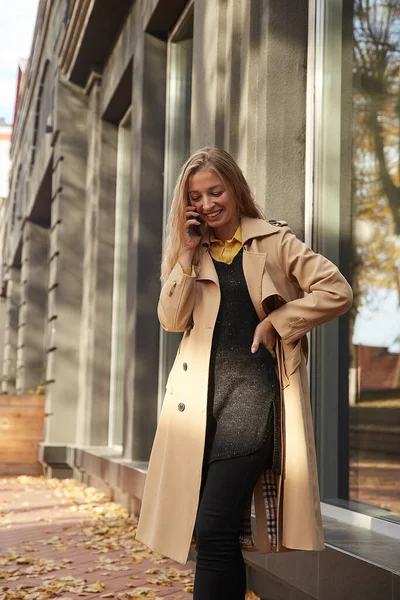 Kaukasisk kvinna i beige trenchcoat ringer via smartphone utomhus på solig dag — Stockfoto