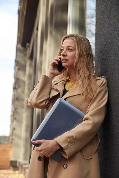 Kaukasiska kvinna i snygg beige päls ringer via smartphone, håller bärbar dator — Stockfoto