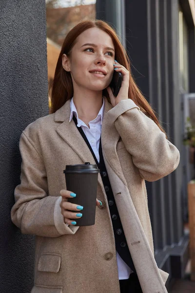 Kaukasiska kvinna i elegant beige rock ringer via smartphone med kopp kaffe — Stockfoto