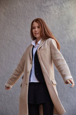 Güzel, beyaz, kızıl saçlı, şık bej ceketli, duvarın önünde.