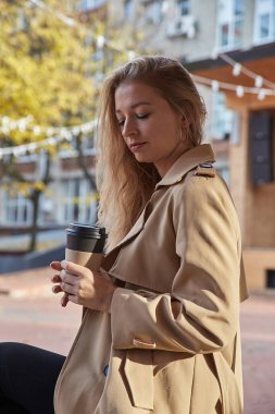 Açık havada bej renkli trençkotlu beyaz kadın portresi ve bir fincan kahve.