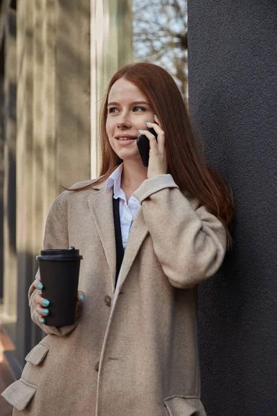 Mujer caucásica en elegante abrigo beige llamando por teléfono inteligente con taza de café —  Fotos de Stock