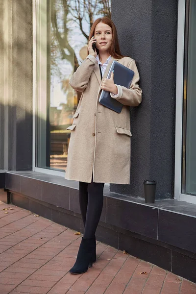 caucasian woman in stylish beige coat calling by smartphone, holding laptop