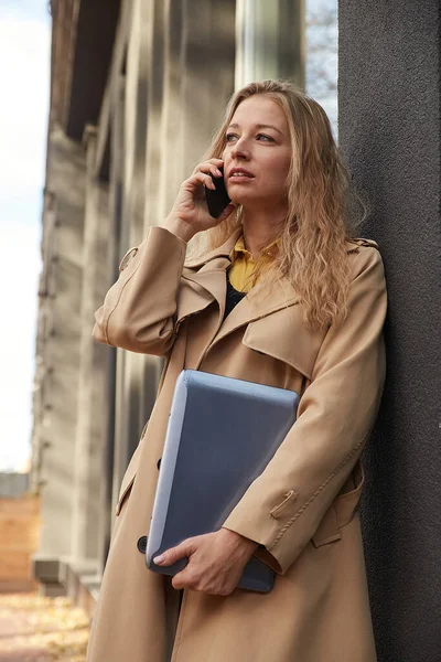 Kaukasiska kvinna i snygg beige päls ringer via smartphone, håller bärbar dator — Stockfoto