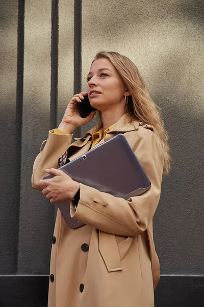 Kaukázusi nő stílusos bézs kabát hívás okostelefonnal, gazdaság laptop — Stock Fotó