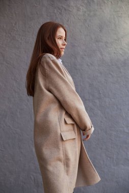 Güzel, beyaz, kızıl saçlı, şık bej ceketli, duvarın önünde.