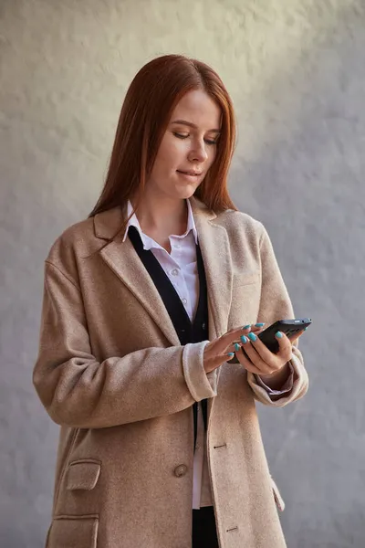 Νεαρή καυκάσια όμορφη κοπέλα σε παλτό χρησιμοποιώντας smartphone, ανάγνωση ειδήσεων, μέσα κοινωνικής δικτύωσης — Φωτογραφία Αρχείου