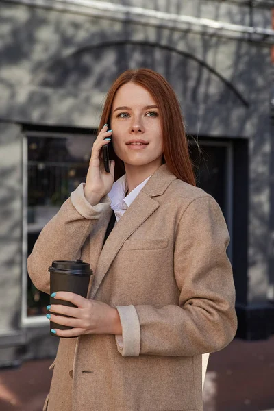 Mujer caucásica en elegante abrigo beige llamando por teléfono inteligente, beber café —  Fotos de Stock