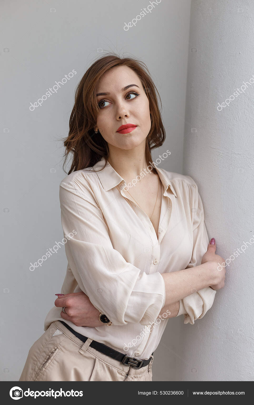 Young stylish beautiful woman wearing pink trousers and silk blouse. Casual  style. Pretty tanned girl with wavy brunette hair standing and posing near  white window and relaxing Stock Photo