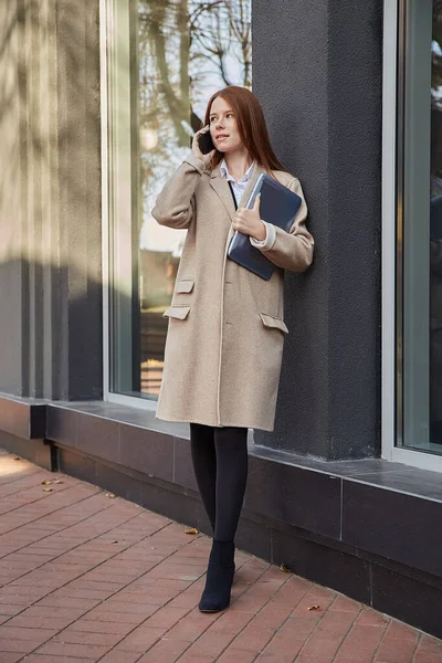 Mulher caucasiana em casaco bege elegante chamando por smartphone, segurando laptop — Fotografia de Stock