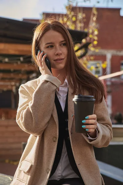Kaukasiska kvinna i snygg beige rock ringer via smartphone, dricka kaffe — Stockfoto