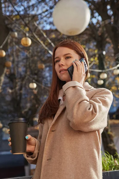 Καυκάσια γυναίκα σε κομψό μπεζ παλτό καλώντας από το smartphone, πίνοντας καφέ — Φωτογραφία Αρχείου