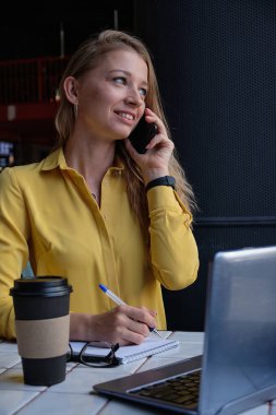 Kafede dizüstü bilgisayarı olan, akıllı telefondan arayan genç ve çekici bir kadın.