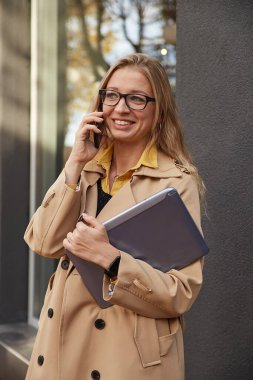 Beyaz kadın şık bej ceketli. Akıllı telefondan arıyor, elinde dizüstü bilgisayar var.