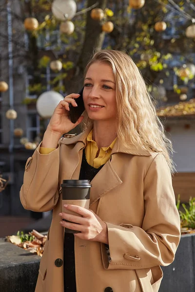Mujer caucásica en elegante abrigo beige llamadas por teléfono inteligente, sostiene la taza de café —  Fotos de Stock