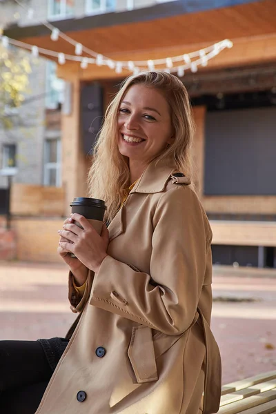 Portræt af hvid kvinde i beige trenchcoat udendørs med kop kaffe Stock-billede