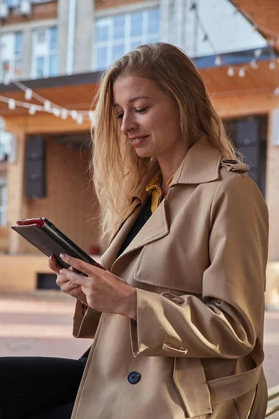 Mujer joven caucásica en abrigo usando tableta al aire libre en el día soleado, leyendo ebook Fotos de stock