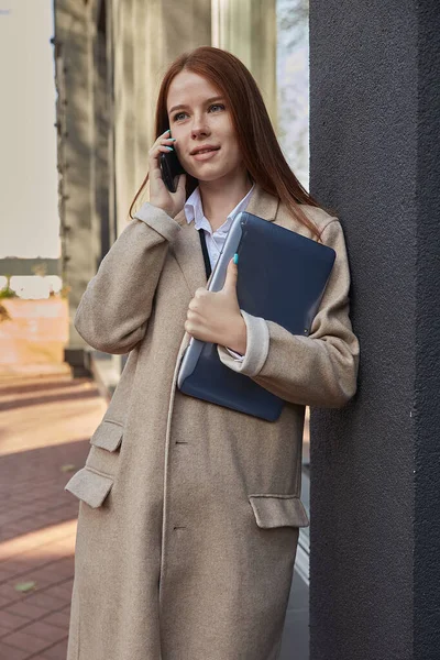Kaukasiske kvinde i stilfuld beige frakke opkald fra smartphone, holder laptop Stock-foto