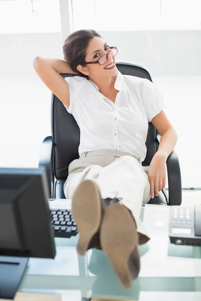 Ontspannen zakenvrouw zit aan haar bureau met haar voeten omhoog glimlachen op camera — Stockfoto