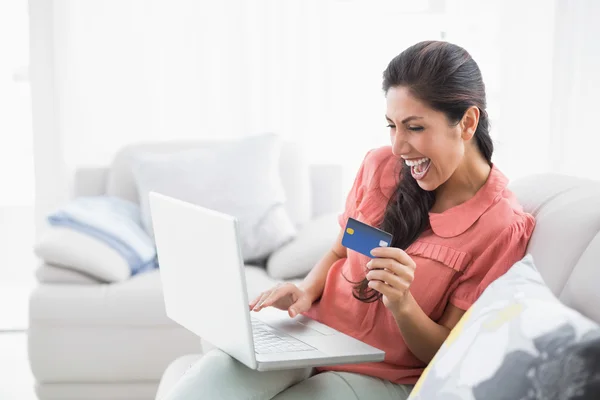 Ecstatic brunette sitting on her sofa using laptop to shop online — Stock Photo, Image