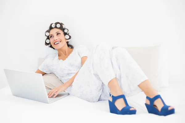 Happy brunette in hair rollers and wedge shoes using her laptop on bed — Stock Photo, Image