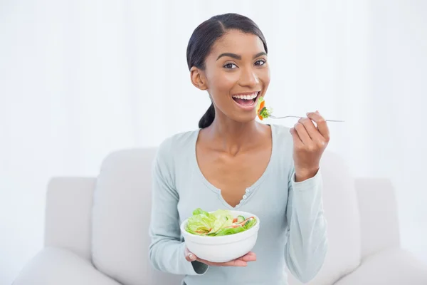 Mujer atractiva sonriente sentada en un sofá acogedor comiendo ensalada — Foto de Stock