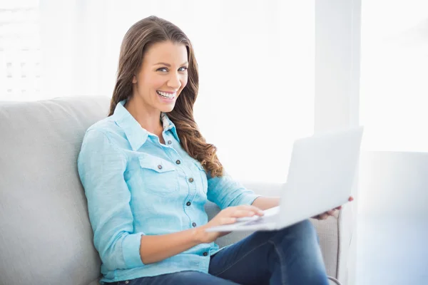 Happy brunette sitting on sofa using laptop — Stock Photo, Image