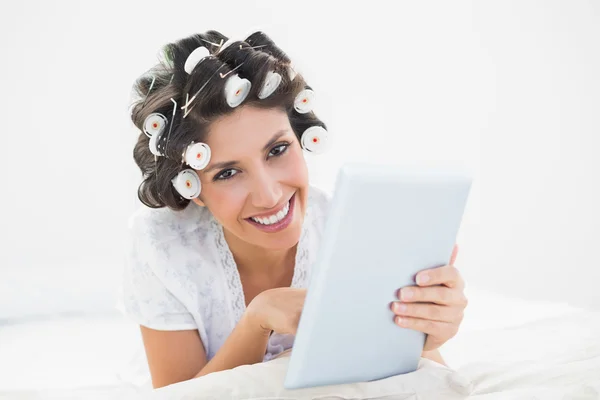 Pretty brunette in hair rollers lying on her bed using her tablet smiling at camera — Stock Photo, Image