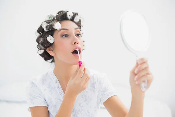 Hübsche Brünette in Haarrollen hält Handspiegel und trägt Lipgloss auf — Stockfoto