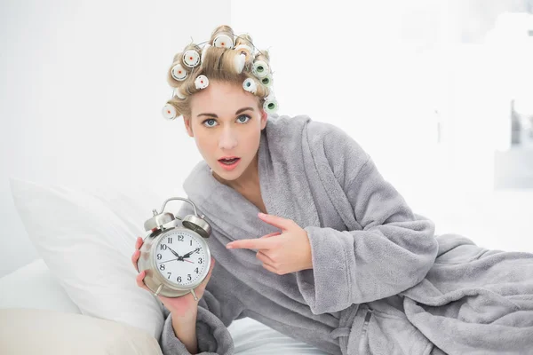 Mulher loira surpreendida em encrespadores de cabelo apontando seu despertador — Fotografia de Stock