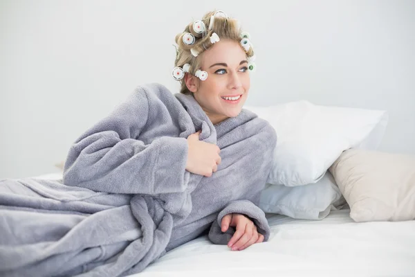 Sonriente mujer rubia relajada en rulos de pelo posando acostada en su cama —  Fotos de Stock