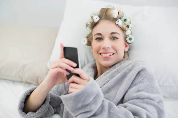 Mulher loira relaxada encantadora em encrespadores de cabelo usando seu telefone móvel — Fotografia de Stock