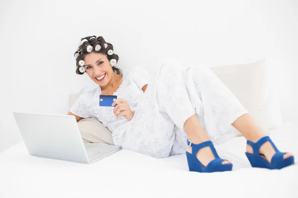 Morena em rolos de cabelo e sapatos cunha usando seu laptop para fazer compras on-line na cama — Fotografia de Stock
