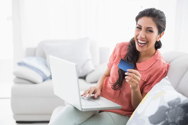Śmiejąc się, brunetka siedzi na jej kanapie przy pomocy laptopa do sklepu online — Zdjęcie stockowe