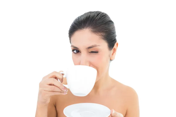 Pretty brunette holding a cup and saucer and taking a sip while winking — Stock Photo, Image